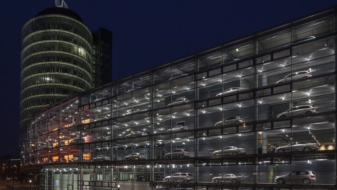 Nighttime exterior of a multi-storey glass Mercedes-Benz dealership with illuminated floors of parked cars