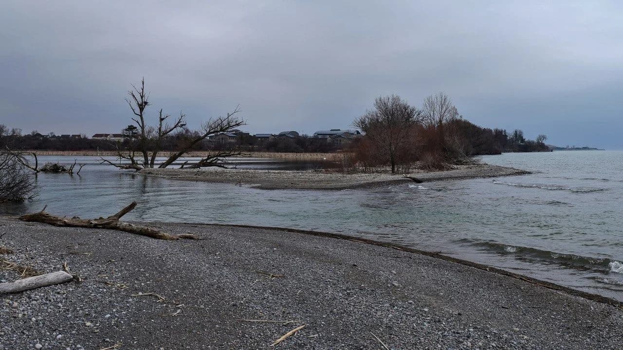 Lynde Creek emptying into Lake Ontario in Whitby, Ontario, with a gravel shoreline and bare trees under a grey sky; no people visible