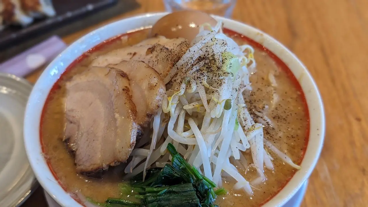 A bowl of ramen topped with sliced pork, a boiled egg, bean sprouts and greens on a wooden table.